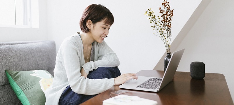 Junge Frau sitzt lächelnd in hellem, weiss, grau, grünem Wohnzimmer am Laptop.