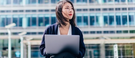 A young businesswoman sits in front of an office complex on a small wall with a laptop on her lap. She looks to the right as if she were looking into the future as a freelancer.