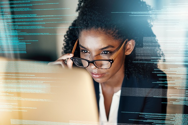 An IT worker with glasses is sitting in front of a computer, IT code can be seen to her left and right.