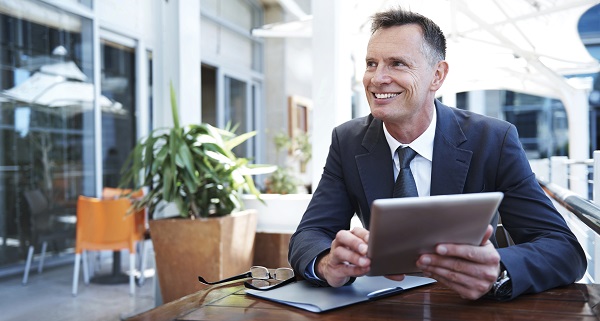 Ein Freelancer im Anzug sitzt mit seinem Laptop in einem Cafe, um von dort aus mobil zu arbeiten. Er lächelt.