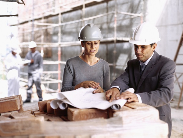 An architect and a construction supervisor go through plans on the construction site. They wear safety helmets and smart clothes.
