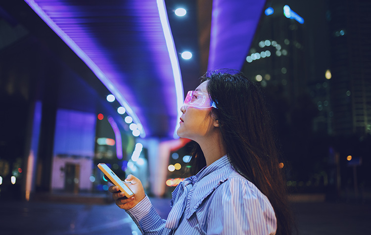 Junge Frau steht mit ihrem Handy und einer Arbeitsbrille unter einer Beleuchtung