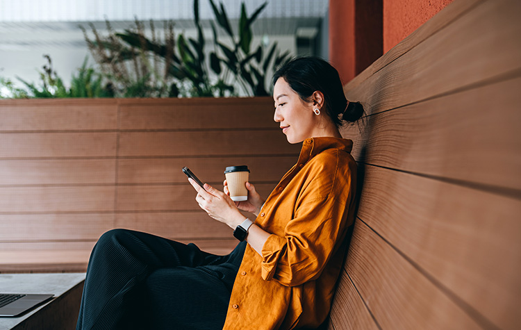 Junge Frau sitzt auf einer Bank, trinkt Kaffee und schaut auf ihr Handy