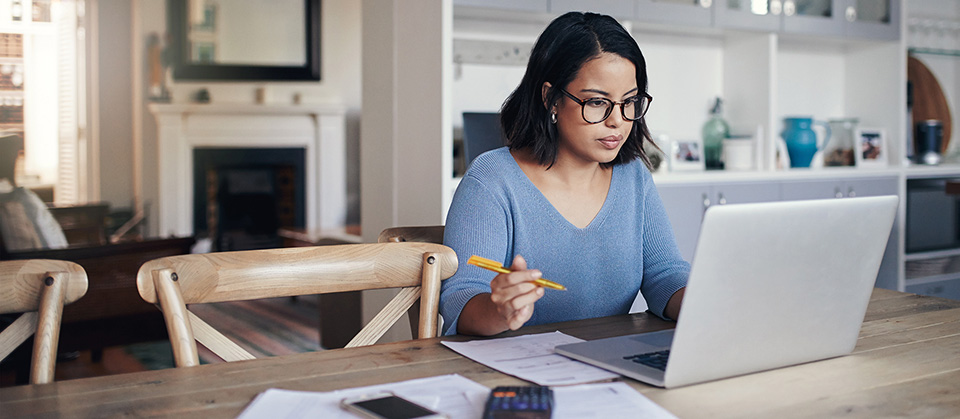 Eine Frau sitzt konzentriert im Homeoffice vor ihrem Laptop. Neben ihr liegen Stifte und Papiere auf einem Tisch aus schickem Massivholz.