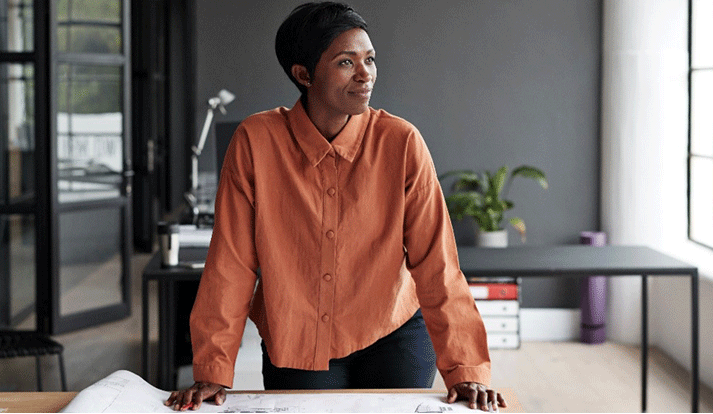 Dynamische Frau steht vor ihrem Schreibtisch mit ausgebreiteten Plänen