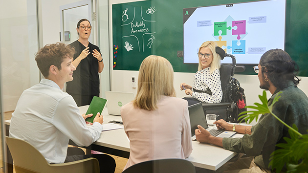 Foto im Besprechungsraum: Eine Person im Rollstuhl präsentiert vor einer Gruppe von vier Personen, während daneben eine weitere Person in Gebärdensprache übersetzt. Im Hintergrund ist ein Bildschirm mit einem Bild zu inklusiver Unternehmenskultur zu sehen.