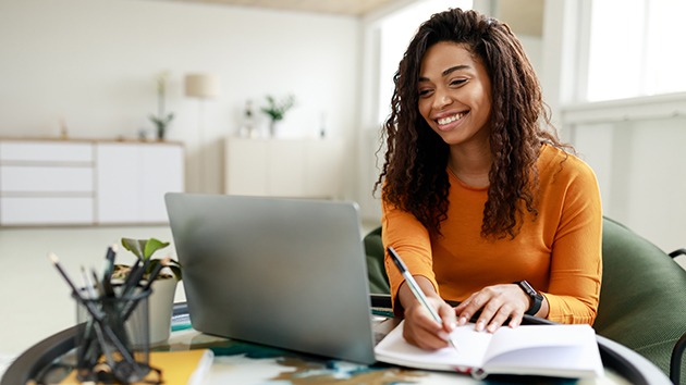 Junge Frau sitzt lächelnd vor ihrem Laptop und macht sich Notizen