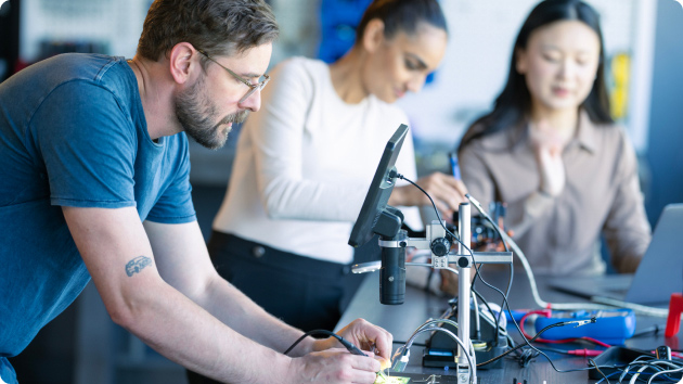 Mann arbeitet mit zwei Kolleginnen an einem Werktisch und lötet an Platinen