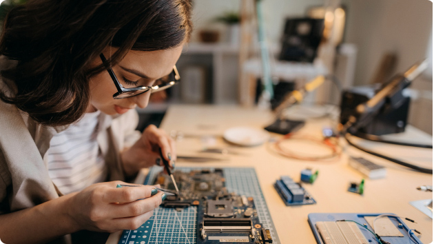 Frau mit Brille lötet an Platine auf einem Tisch 