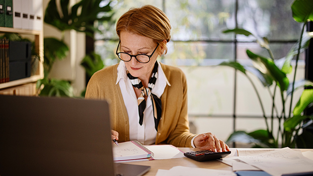 Frau sitzt am Schreibtisch und rechnet mit einem Taschenrechner
