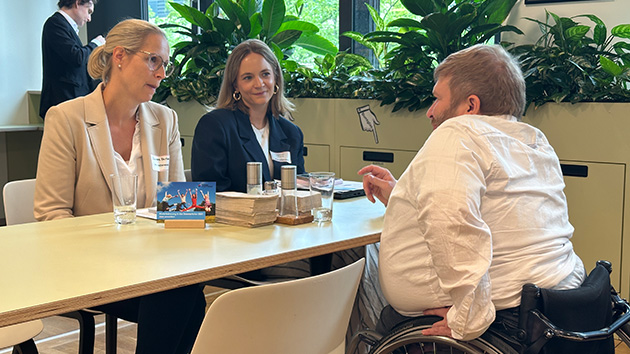 Foto Bewerbungssituation: 3 Personen in Business-Kleidung sitzen an einem Tisch. Zwei hinter dem Tisch, die Person vor dem Tisch nutzt einen Rollstuhl. Sie unterhalten sich angeregt. Auf dem Tisch sind Flyer und Wassergläser zu sehen. Im Hintergrund eine Fensterfront mit Pflanzen.