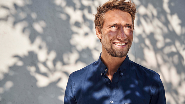 Mann steht lächelnd im Schatten der Blätter eines Baumes